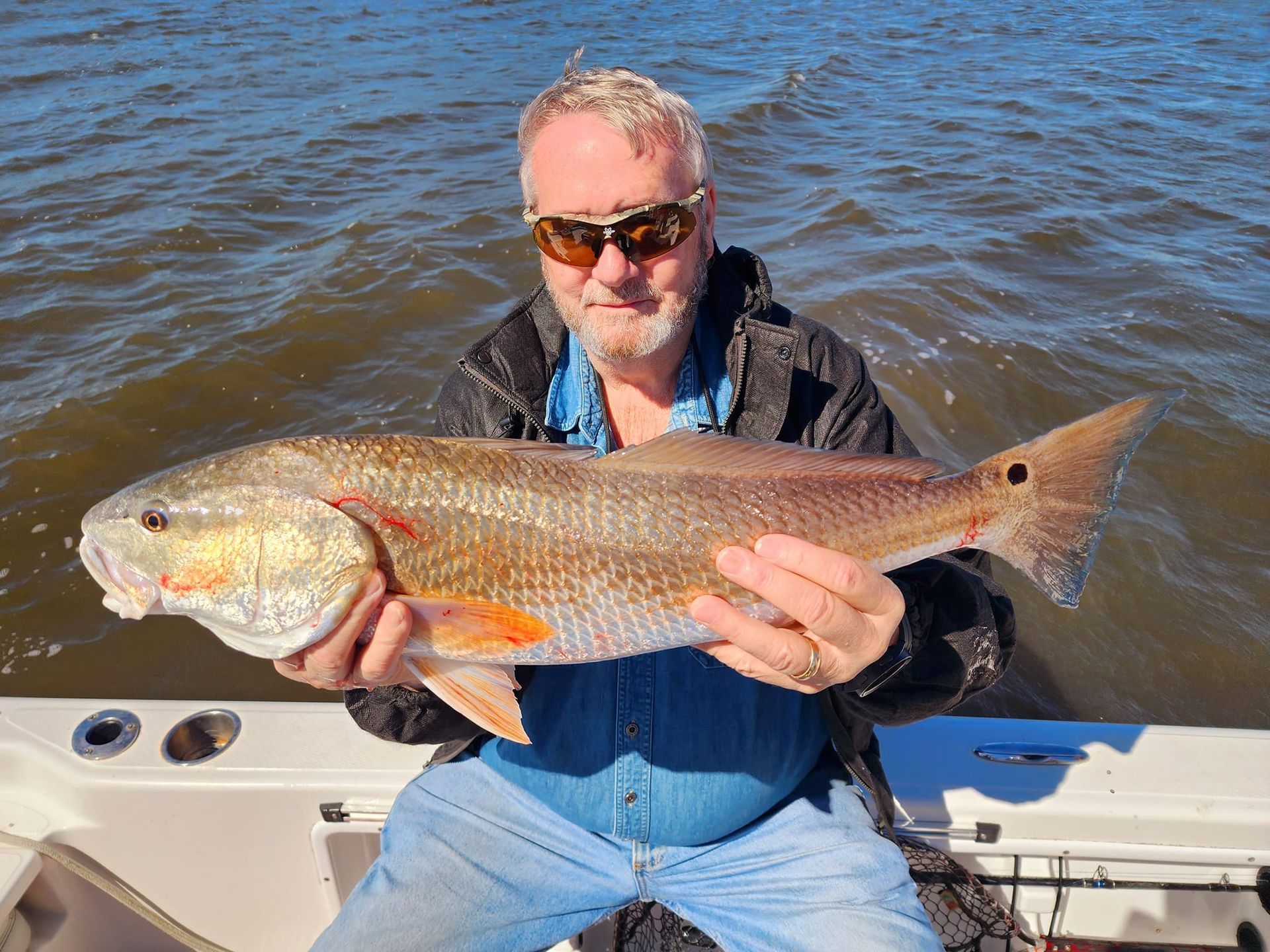 Man holding a large redfish on a boat; water and sky in the background. Fish is reddish-brown and silver.