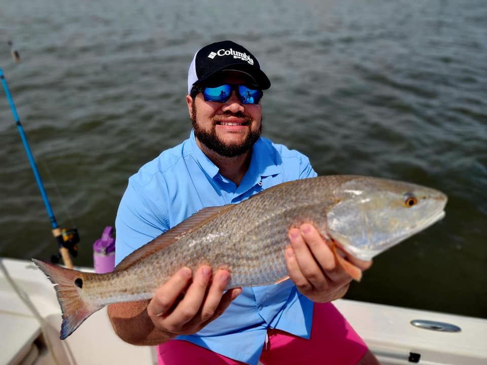 Man on a boat holding a redfish. He's smiling and wearing sunglasses, a blue shirt, and pink shorts.