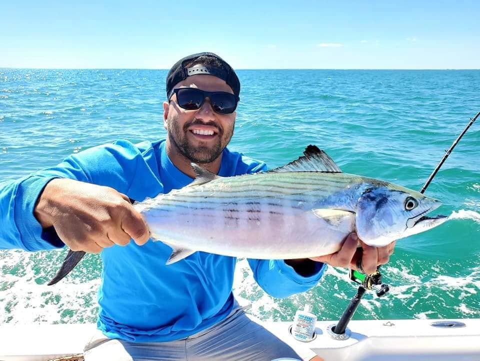 Man on boat smiles, holding a caught fish against a blue ocean backdrop.