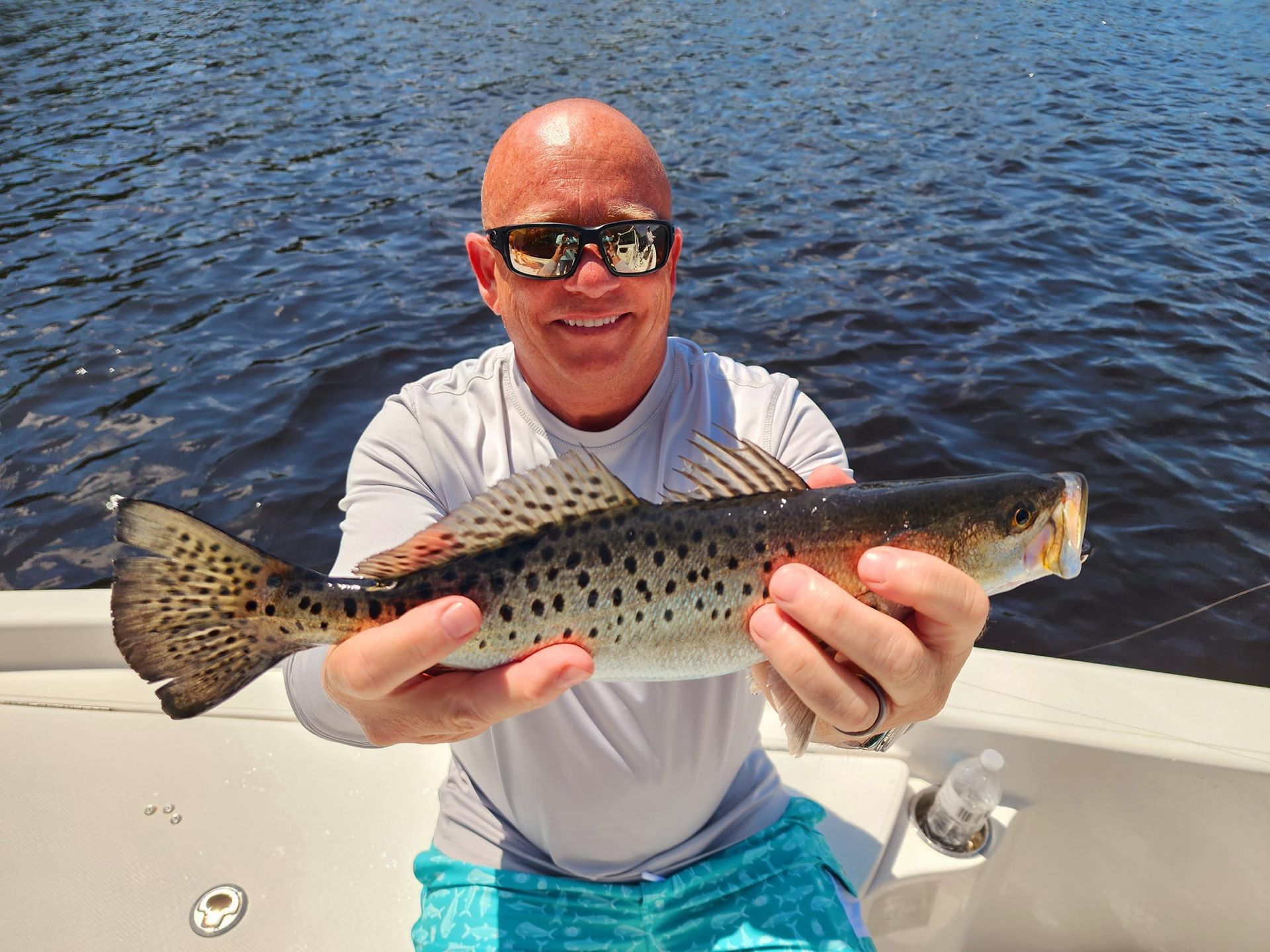 Man holding a speckled trout on a boat, wearing sunglasses and a light blue shirt. Water in the background.