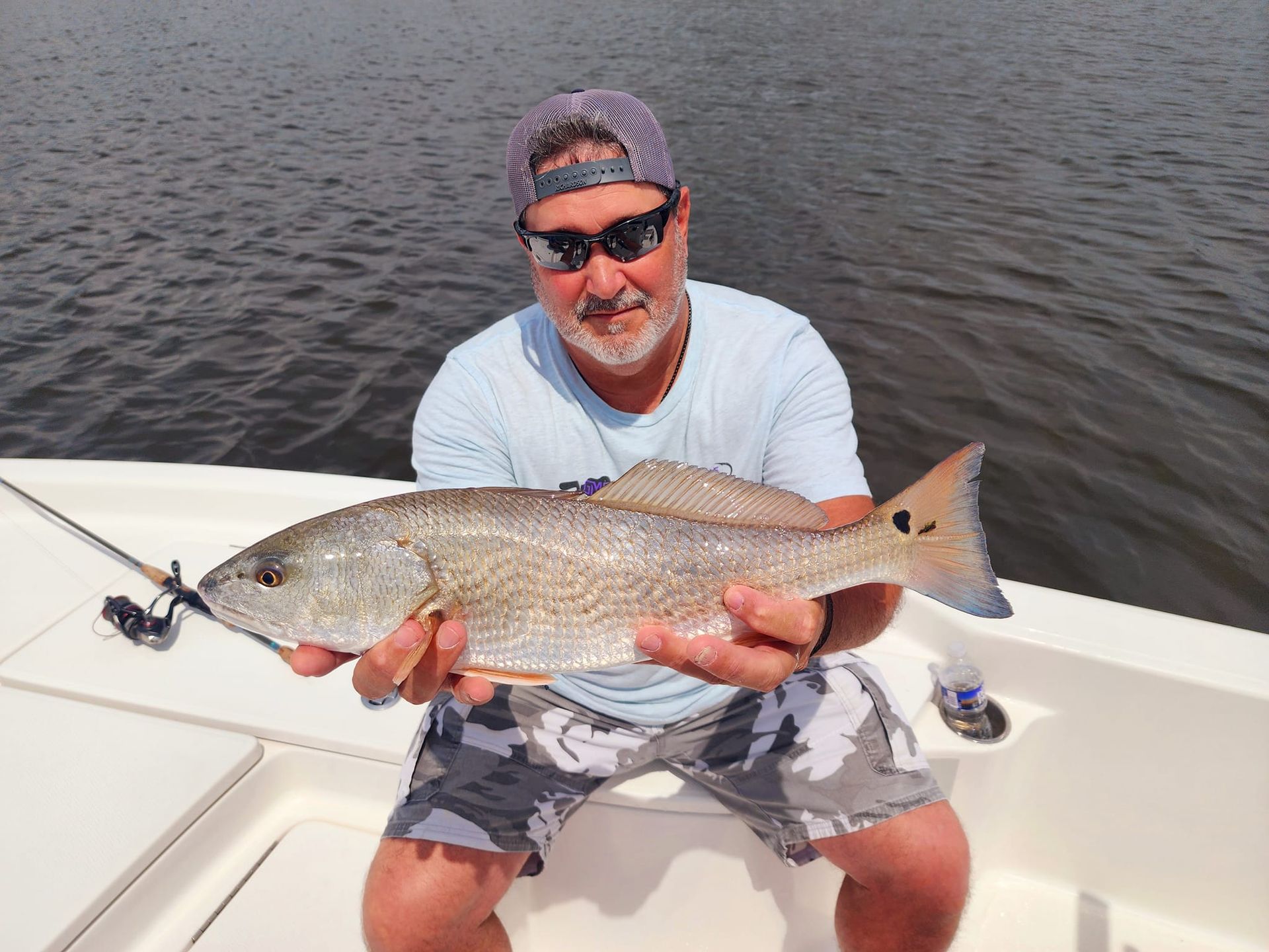 Man holding a redfish on a boat, wearing sunglasses and a hat. Sunny day, water background.
