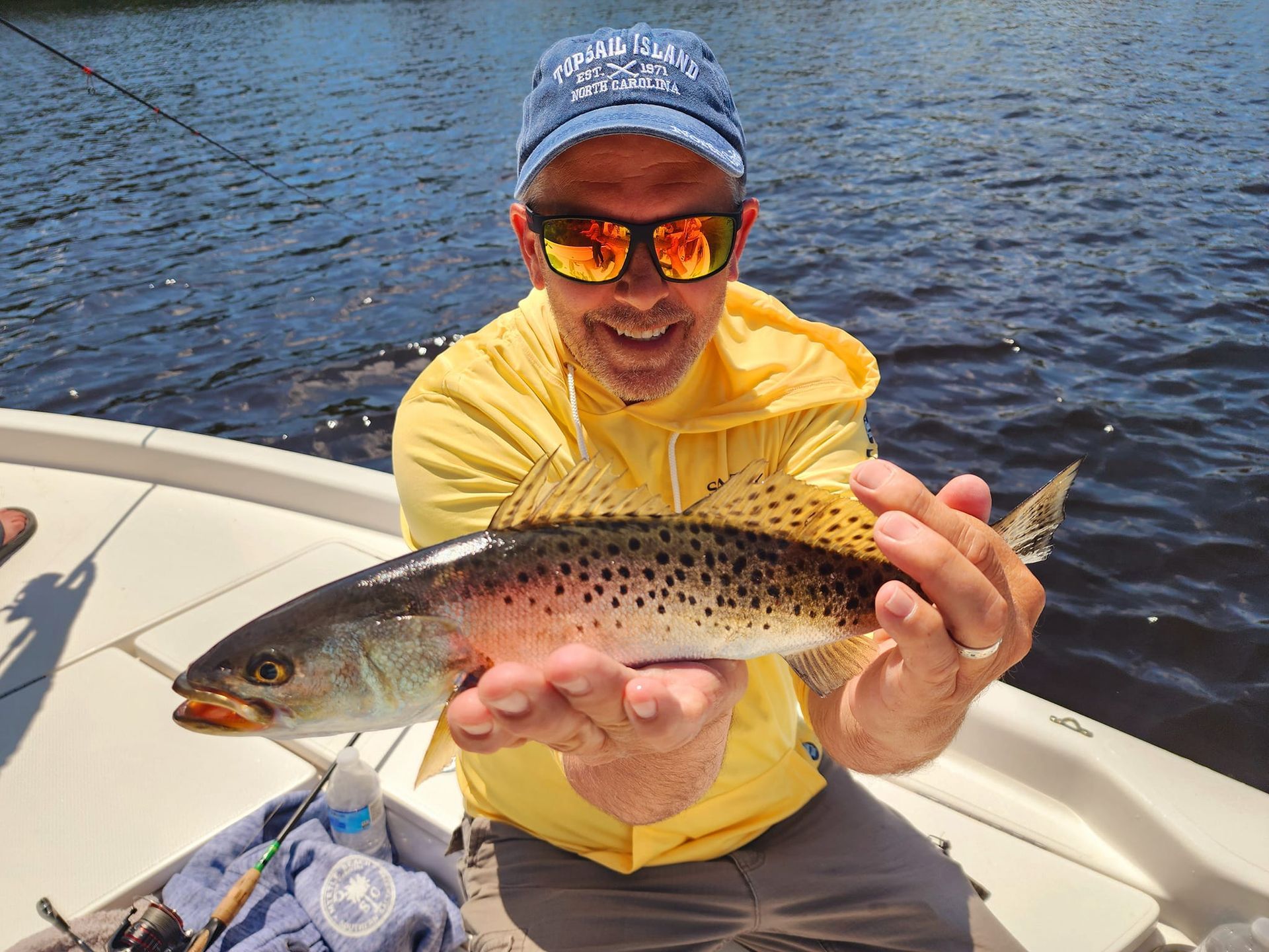 Man holding a speckled trout, smiling, on a boat, wearing sunglasses and a yellow shirt.