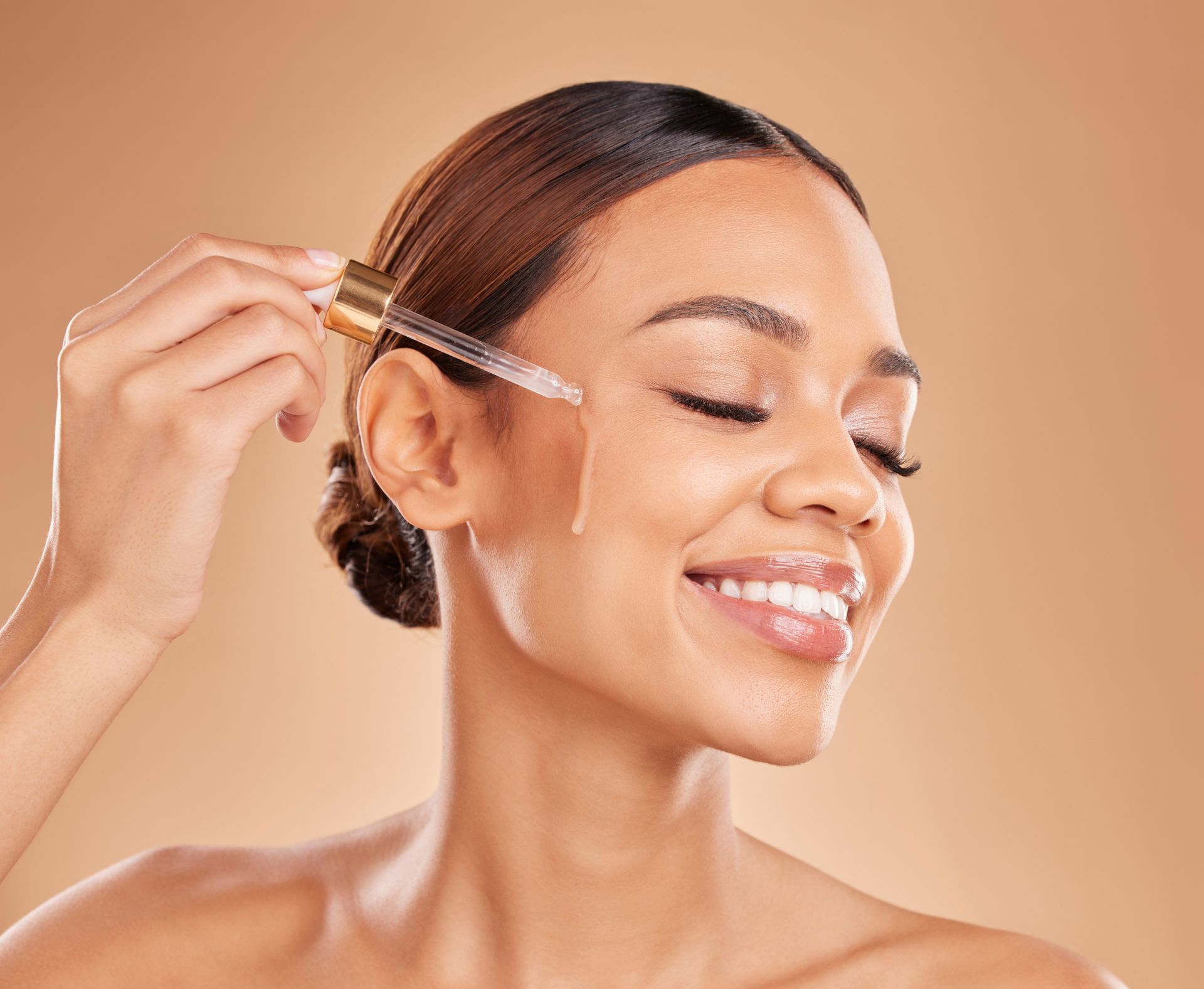 Woman smiling while applying serum with a dropper against a warm beige background