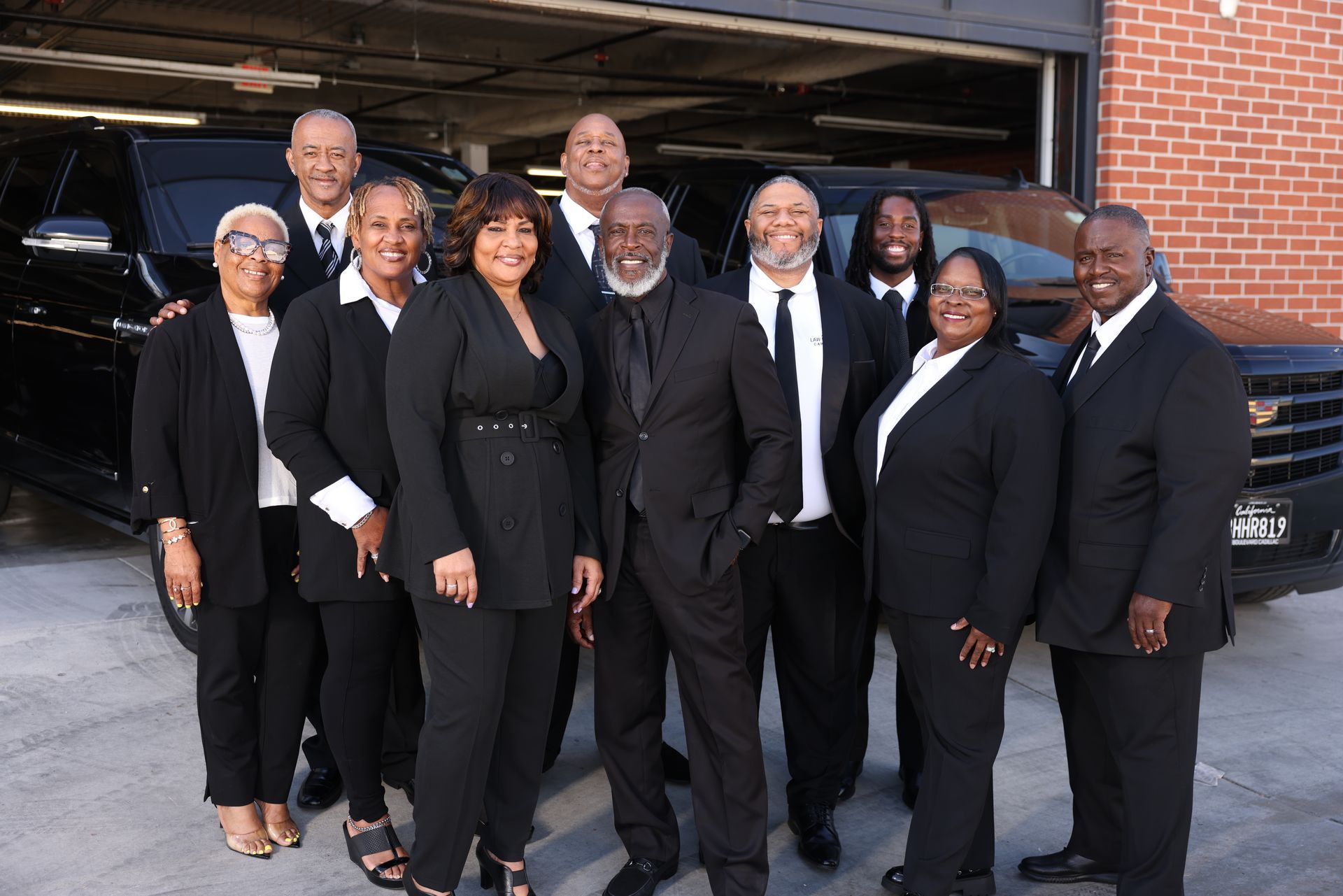A group of ten professionals in formal black suits stand in a row in front of a garage and dark vehicles.