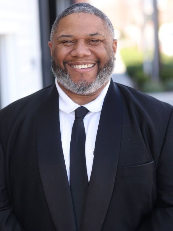 Smiling man in a black suit and tie outdoors with a blurred background