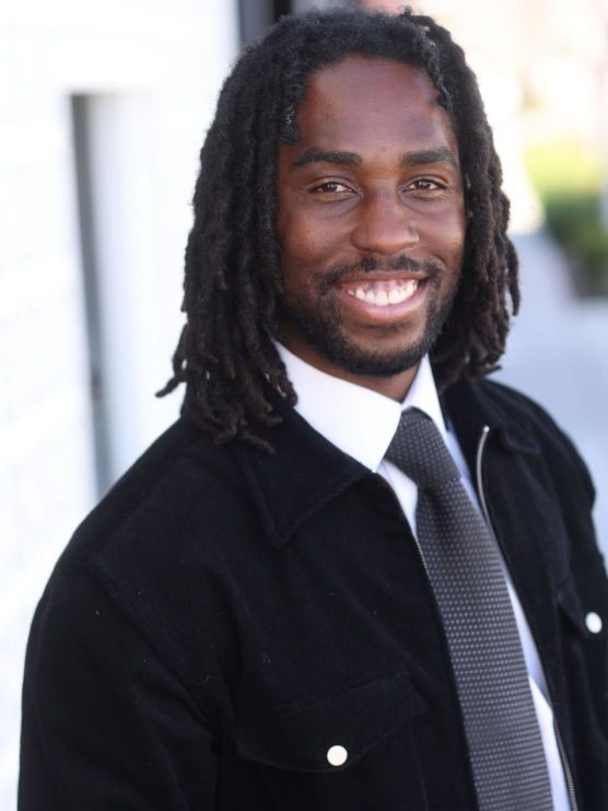 Smiling person in a black jacket and tie against a blurred outdoor background