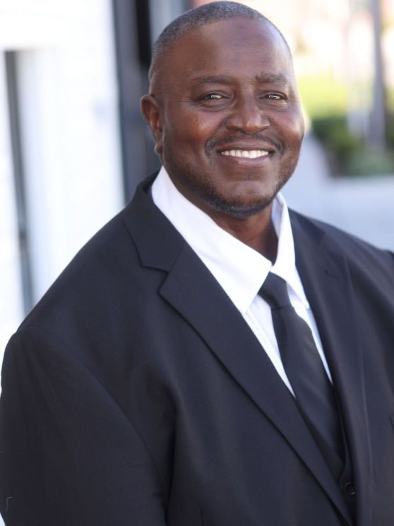 Smiling man in a dark suit and tie, standing indoors against a bright background.