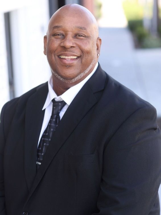Smiling man in a dark suit and tie standing outdoors against a bright background