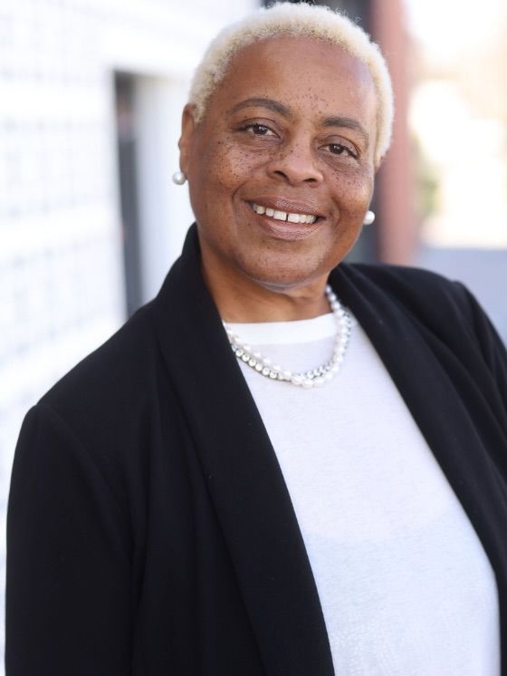 Smiling person with short white hair wearing a black blazer and white top against a softly blurred background