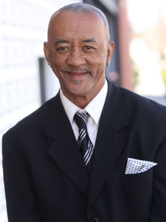 Smiling man in a black suit and striped tie with a pocket square, standing indoors.