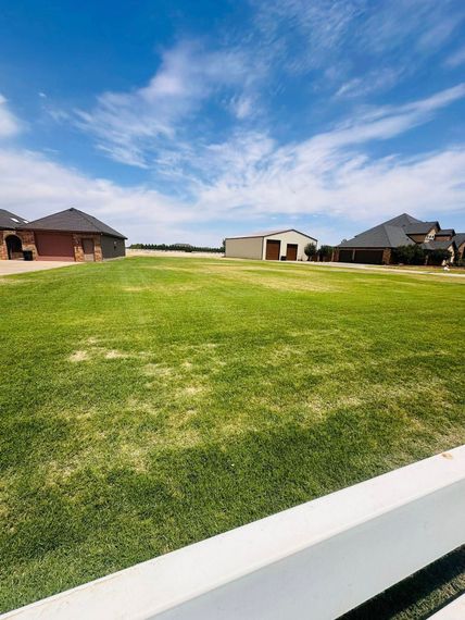 Wide grassy field with scattered houses under a blue, partly cloudy sky, viewed from a white fence