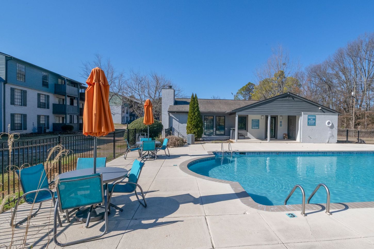 Outdoor apartment pool with lounge chairs, orange umbrellas, and clear blue water under a sunny sky