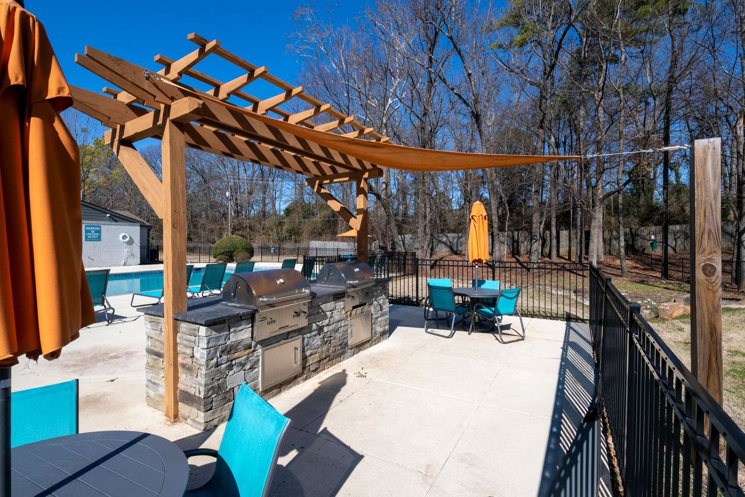 Outdoor patio with turquoise chairs, umbrellas, and a wooden pergola overlooking a pool and trees