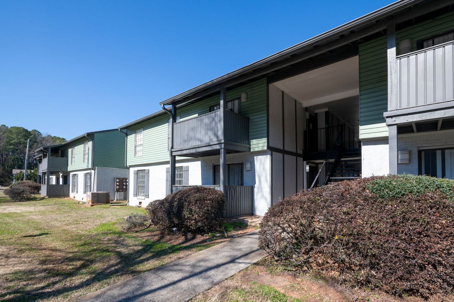 Two-story apartment buildings with green awnings and shrubs along a sunny walkway