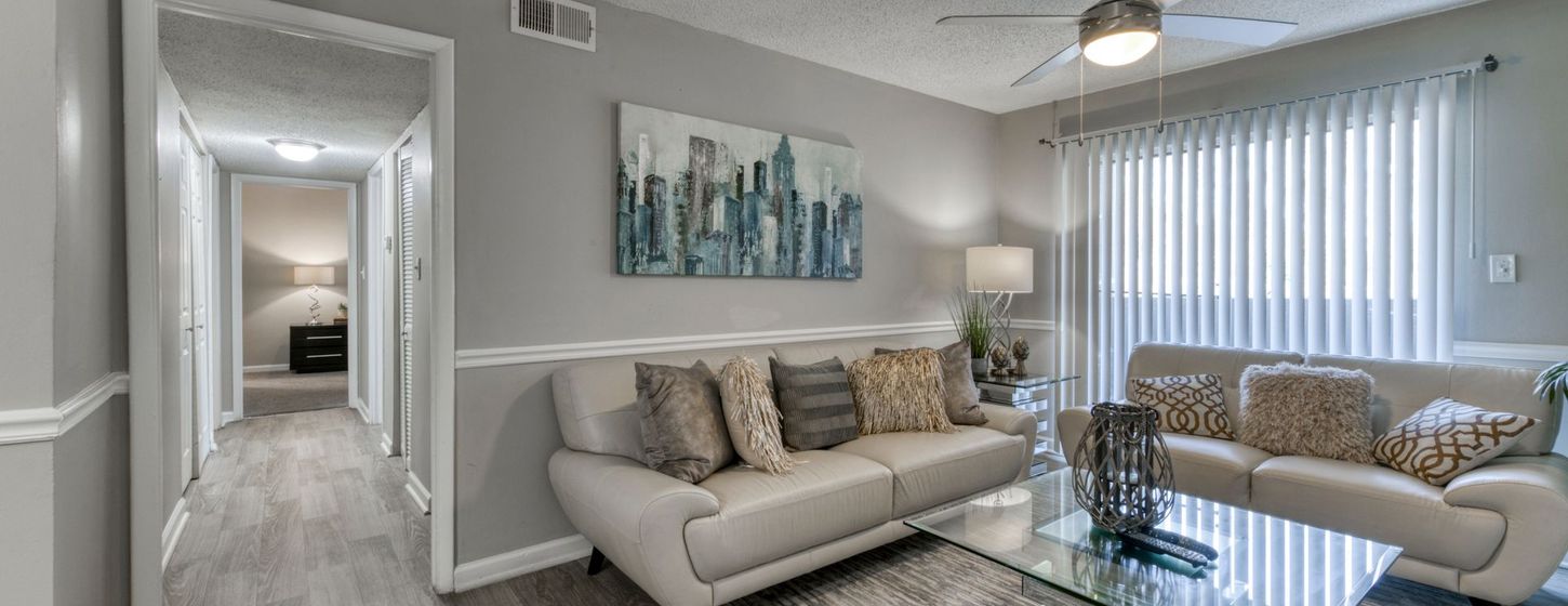 Bright living room with two sofas, glass coffee table, wall art, and hallway view through an open doorway