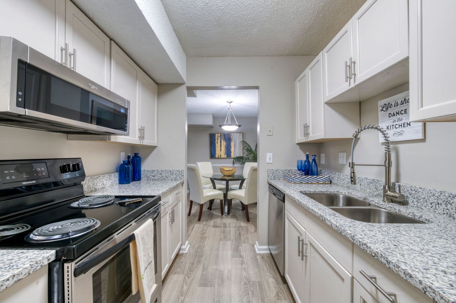 Modern white galley kitchen with black appliances, granite counters, and dining area beyond