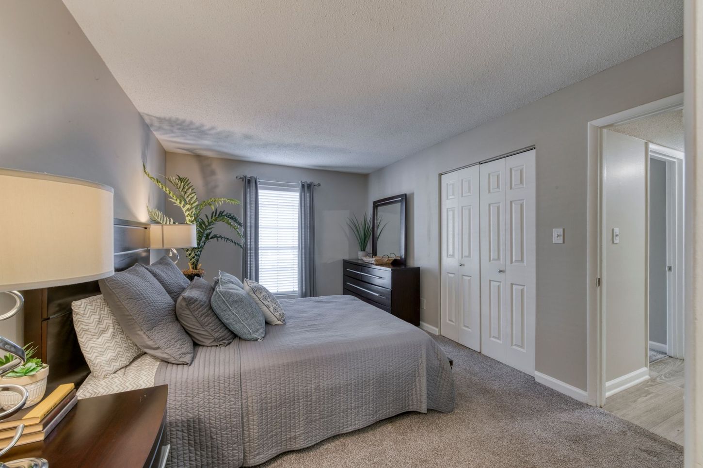 Bright bedroom with a gray bed, beige carpet, dresser, and doorway to a bathroom.