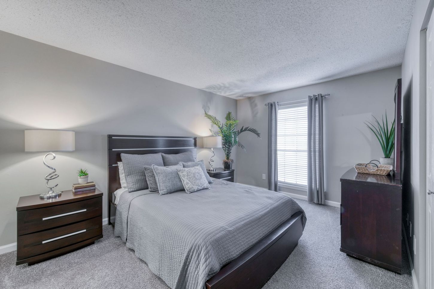 Bright gray bedroom with a bed, dresser, nightstand, and window with curtains.