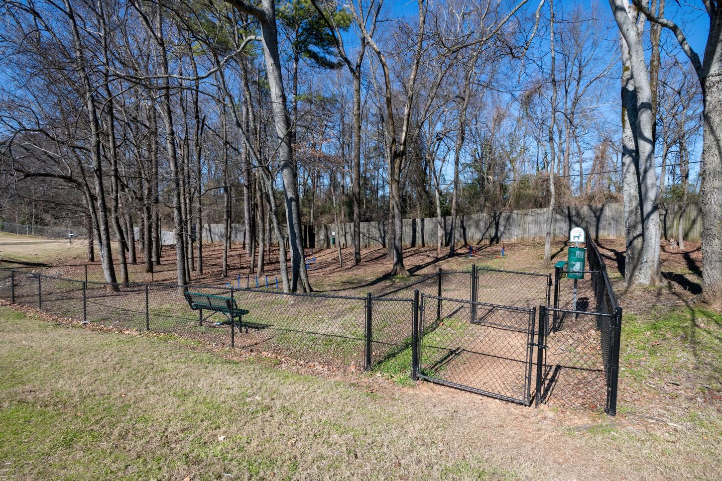 Wooded park with fenced gravel path, benches, and a person in a green jacket standing near the gate.