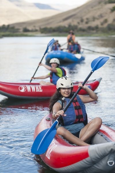 People kayaking on a river, mountains in the background. Smiling kayakers wear helmets and life vests.