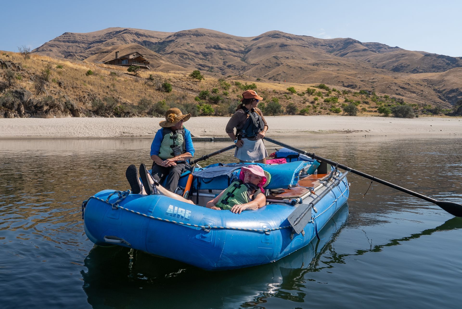 Blue raft with three people on a calm river; mountains in the background. Sunny day.