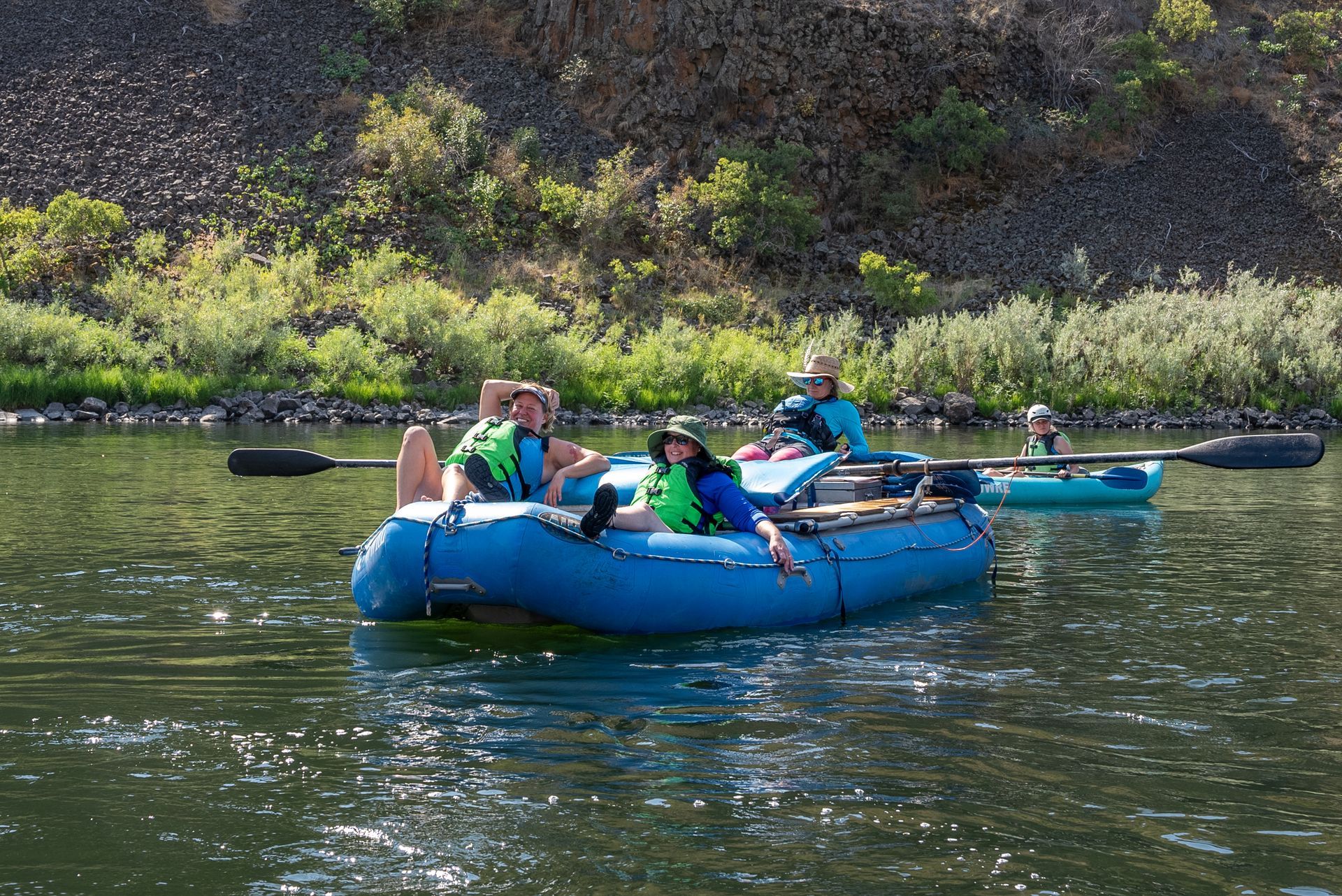 People relaxing on a blue raft on a river, other person paddles a kayak nearby. Rocky bank in the background.