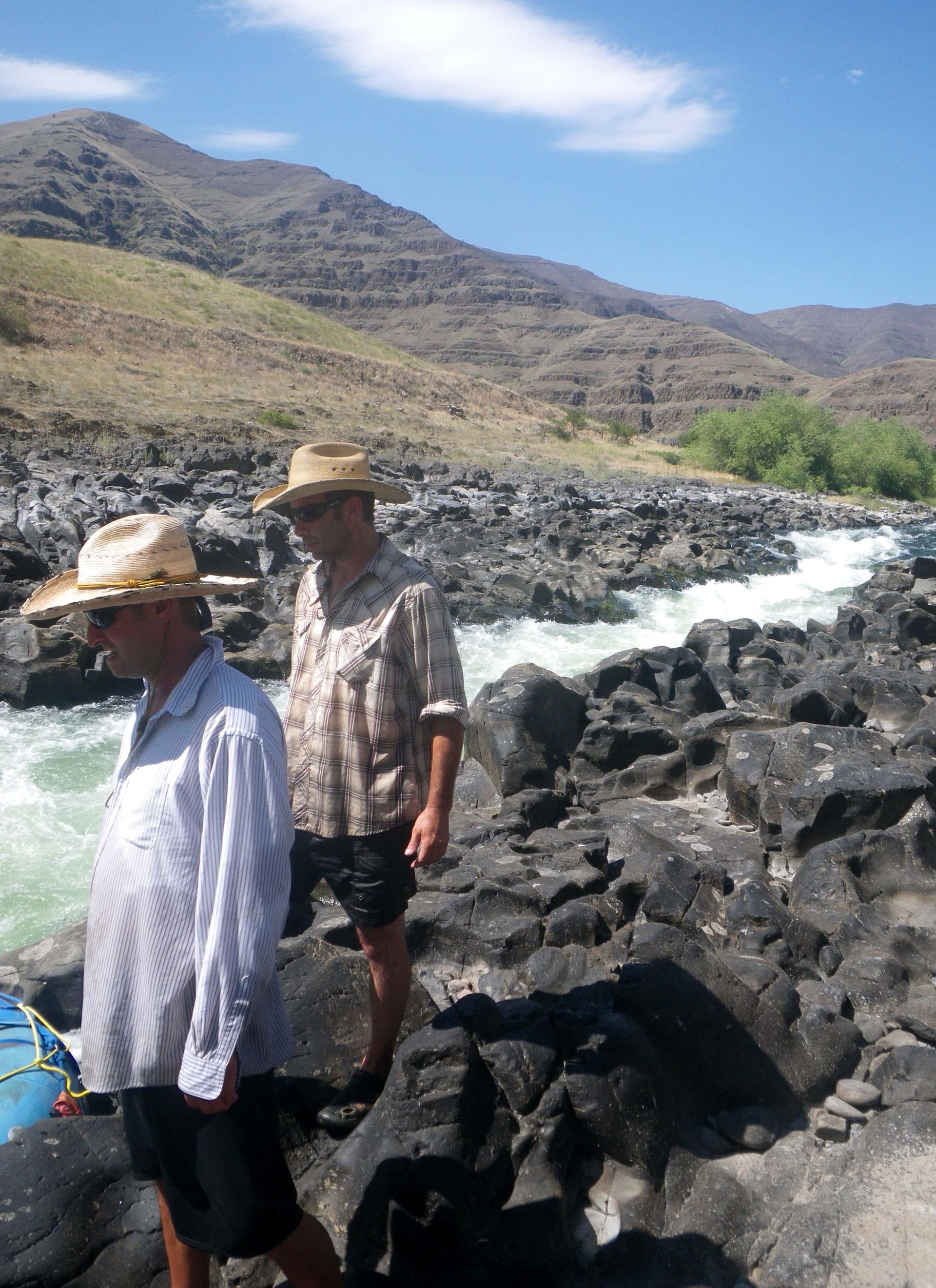 Two men are walking on rocks near a river with mountains in the background