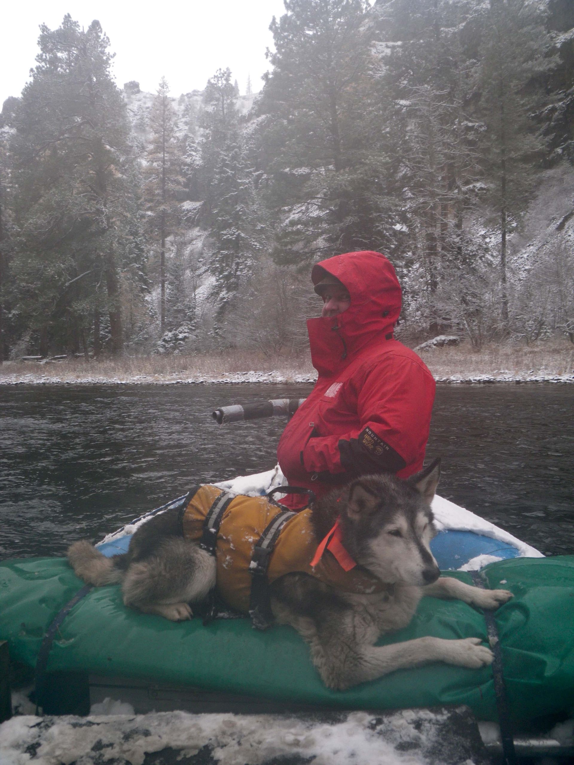 A man in a red jacket is sitting on a green raft with a dog on it.