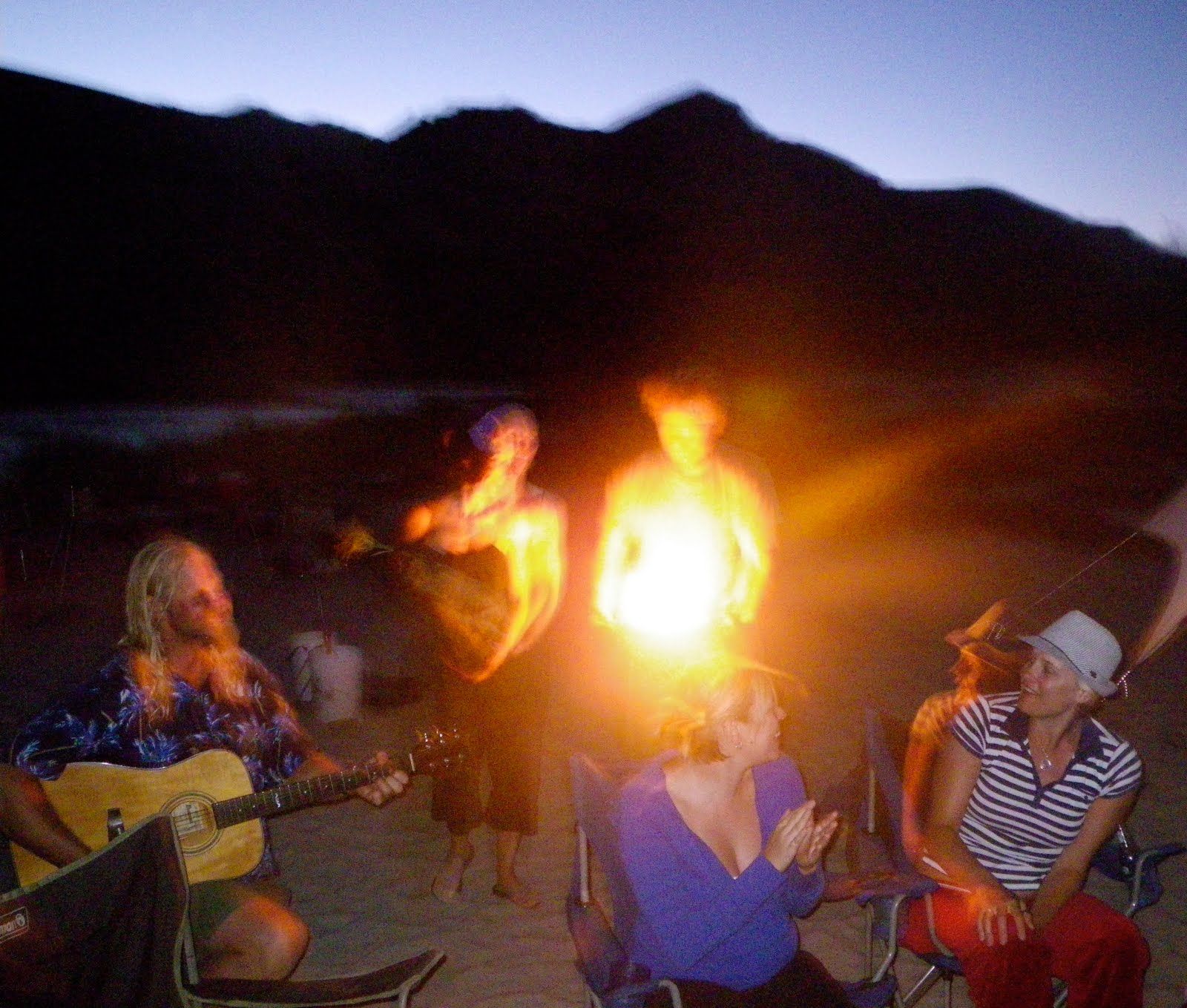 A group of people are gathered around a campfire with a man playing a guitar