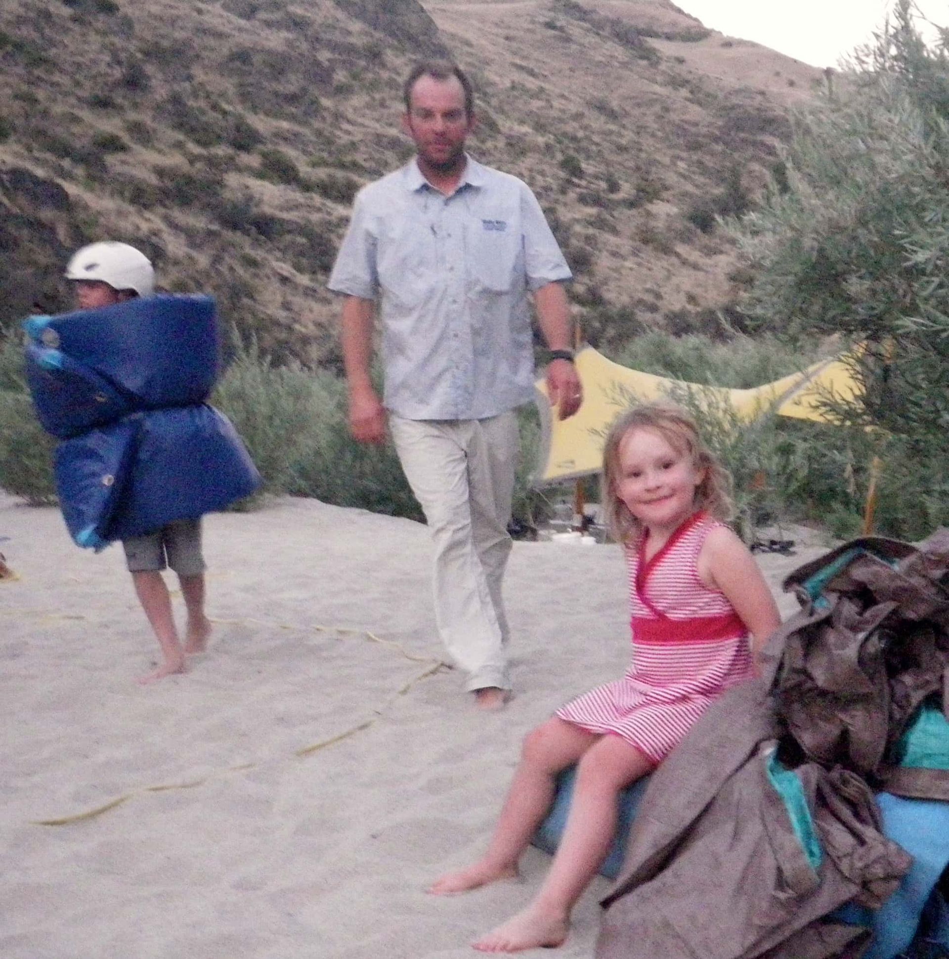 A little girl is sitting on a bag while a man walks behind her
