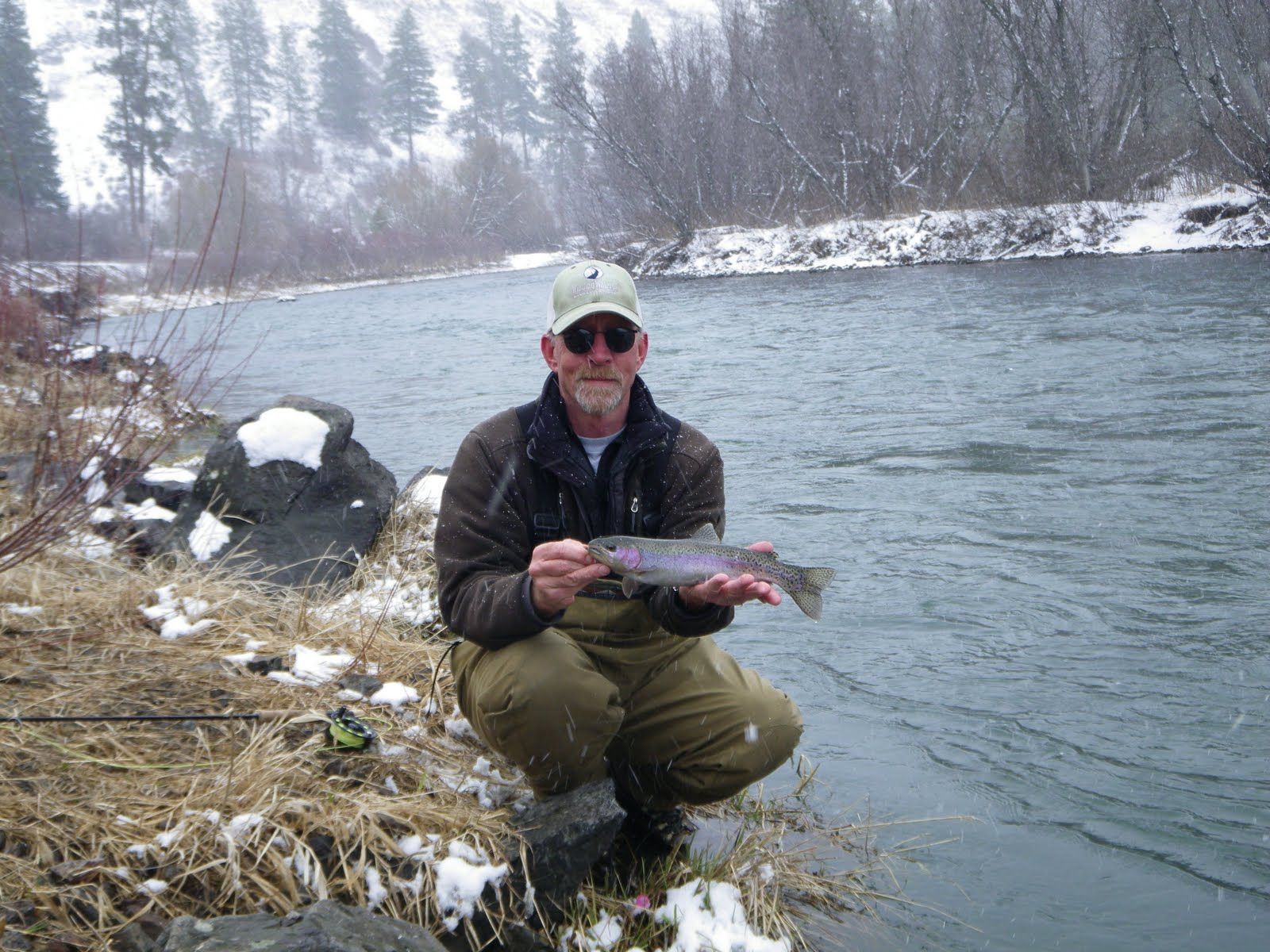 A man is kneeling down next to a river holding a fish.