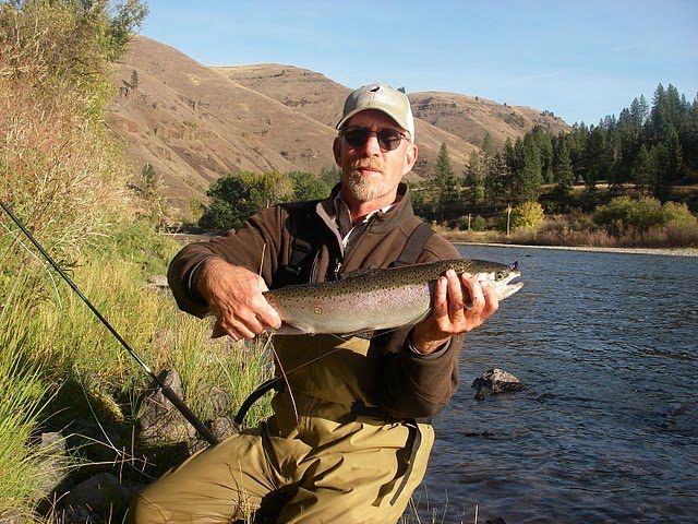 A man is holding a rainbow trout in front of a river
