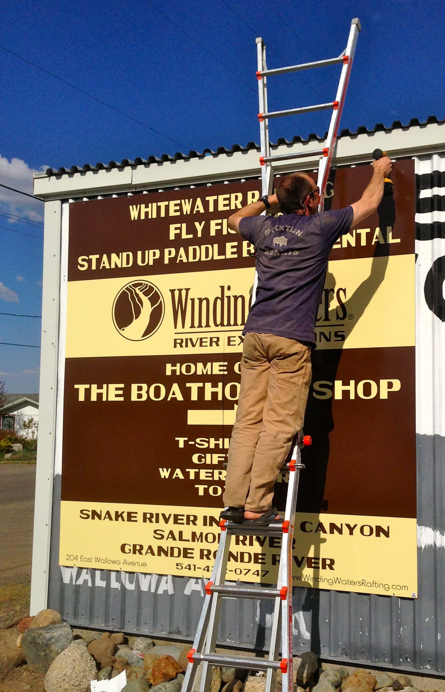 A man on a ladder hanging a sign for the boat shop