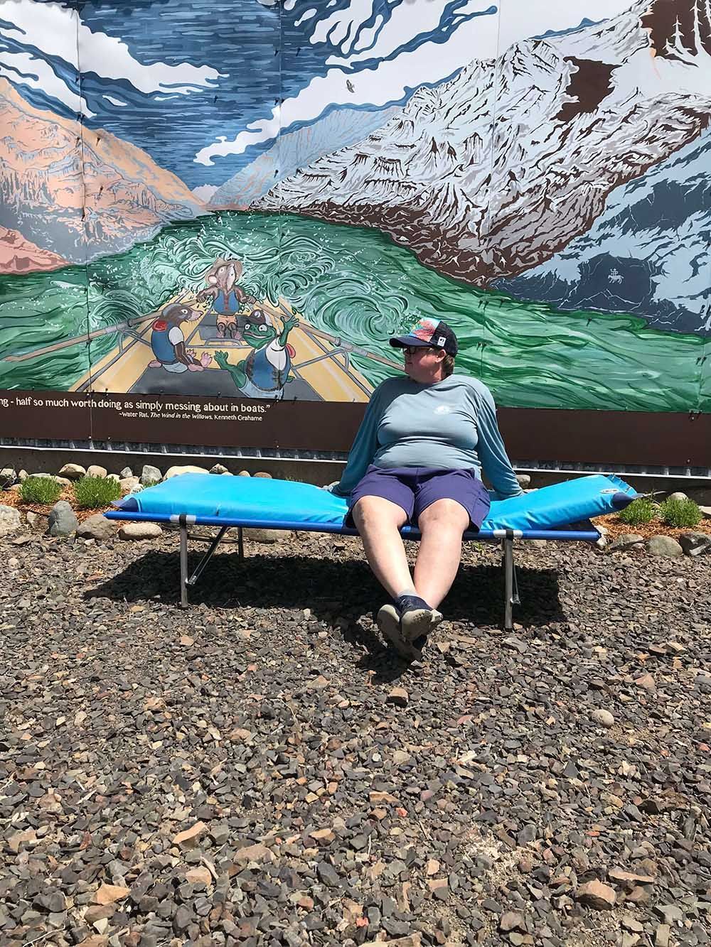 A woman is sitting on a bench in front of a painting of mountains.