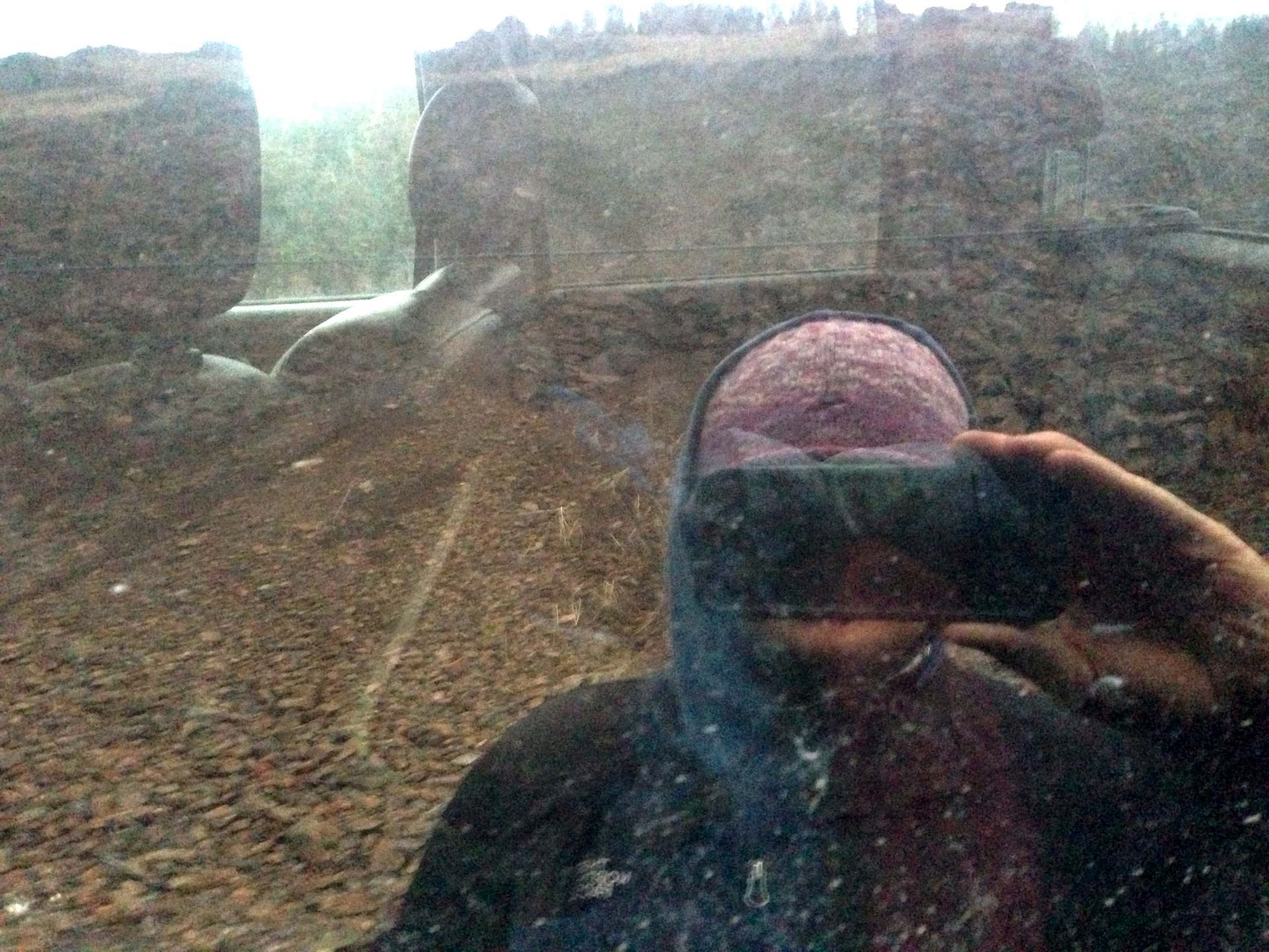 Person in a dark hoodie taking a selfie, blurred outdoor setting with dirt and rocks.