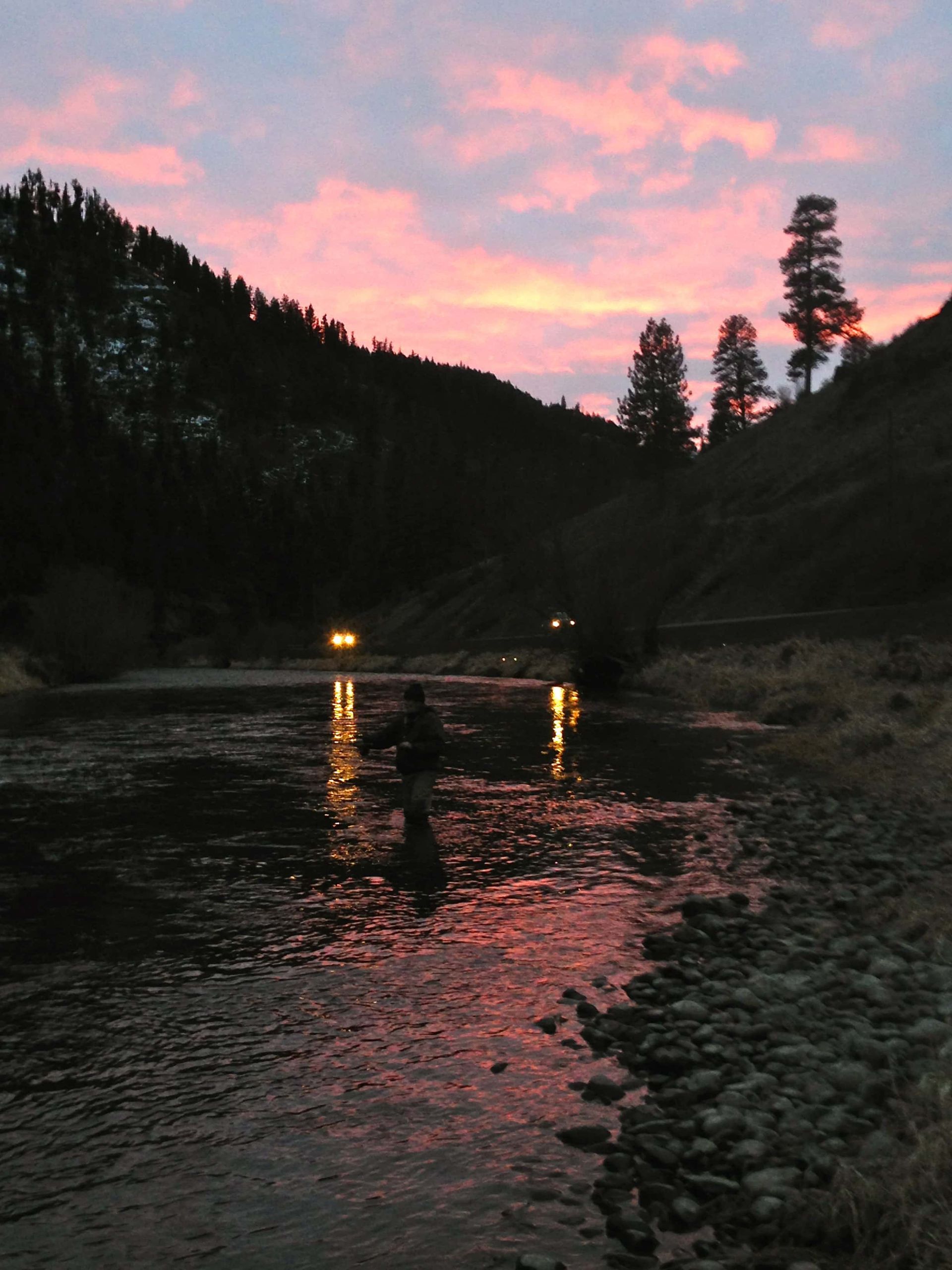 Sunset over a river reflecting pink sky. Two figures fishing, surrounded by dark hills and trees.