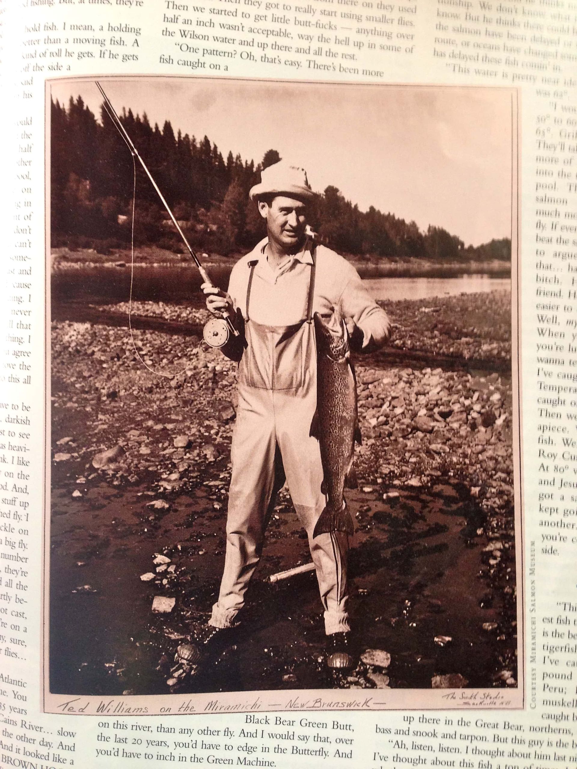 Man in overalls and hat holding a large fish and fishing rod near a river.