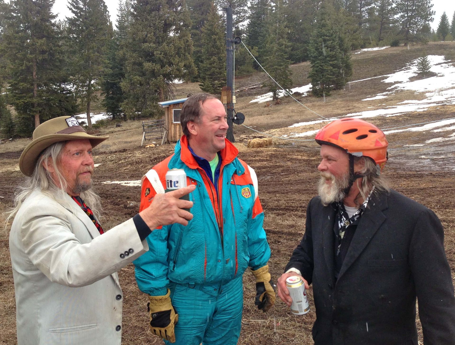 Three men are standing in a field and one of them is wearing an orange helmet