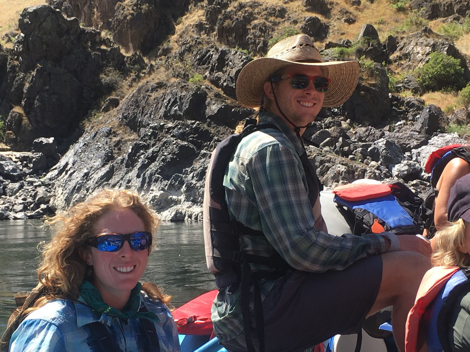 Two people on a raft smile, wearing sunglasses and life vests, against a rocky backdrop.