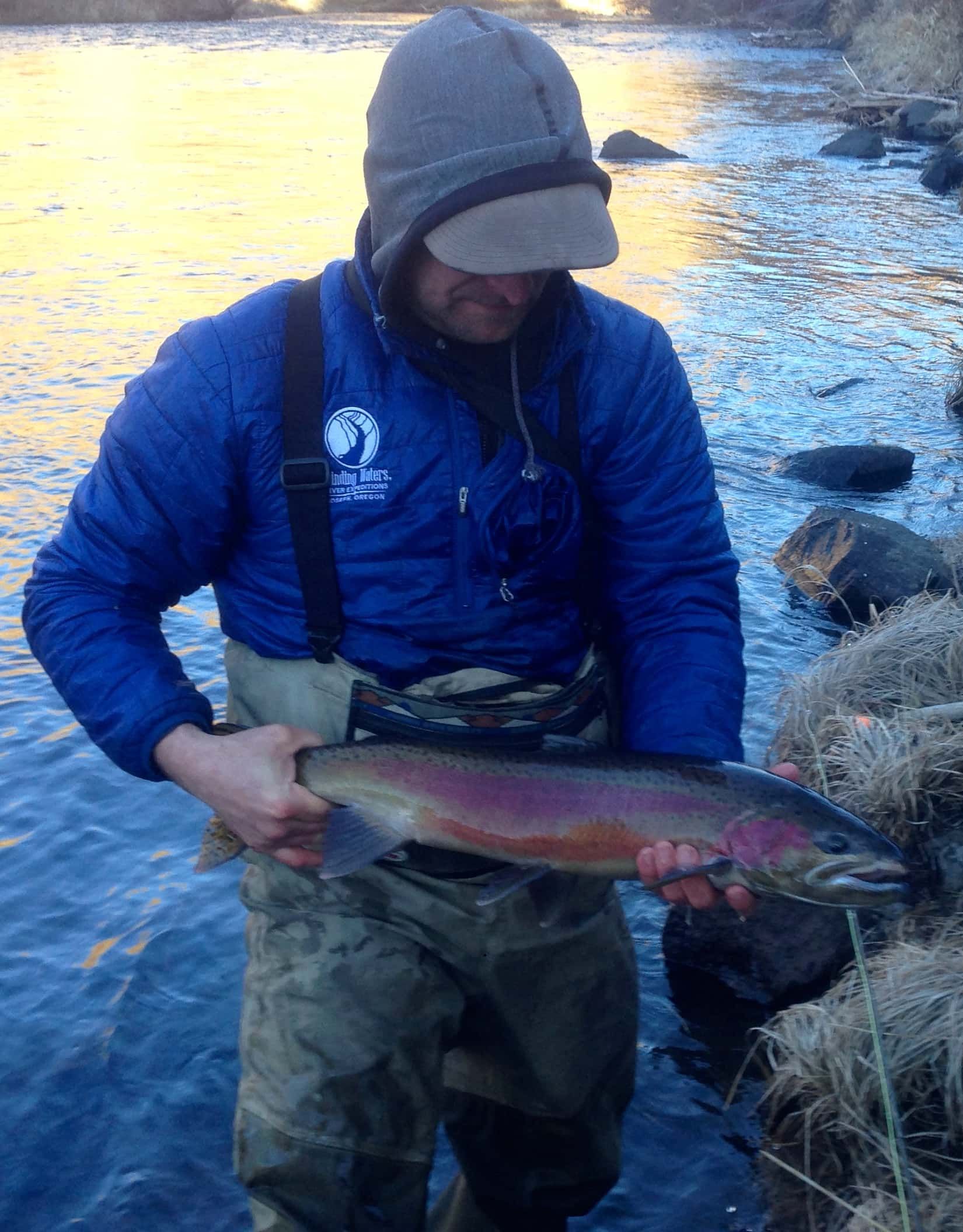Angler holding a large rainbow trout in a river, wearing blue jacket, hooded hat, and waders.