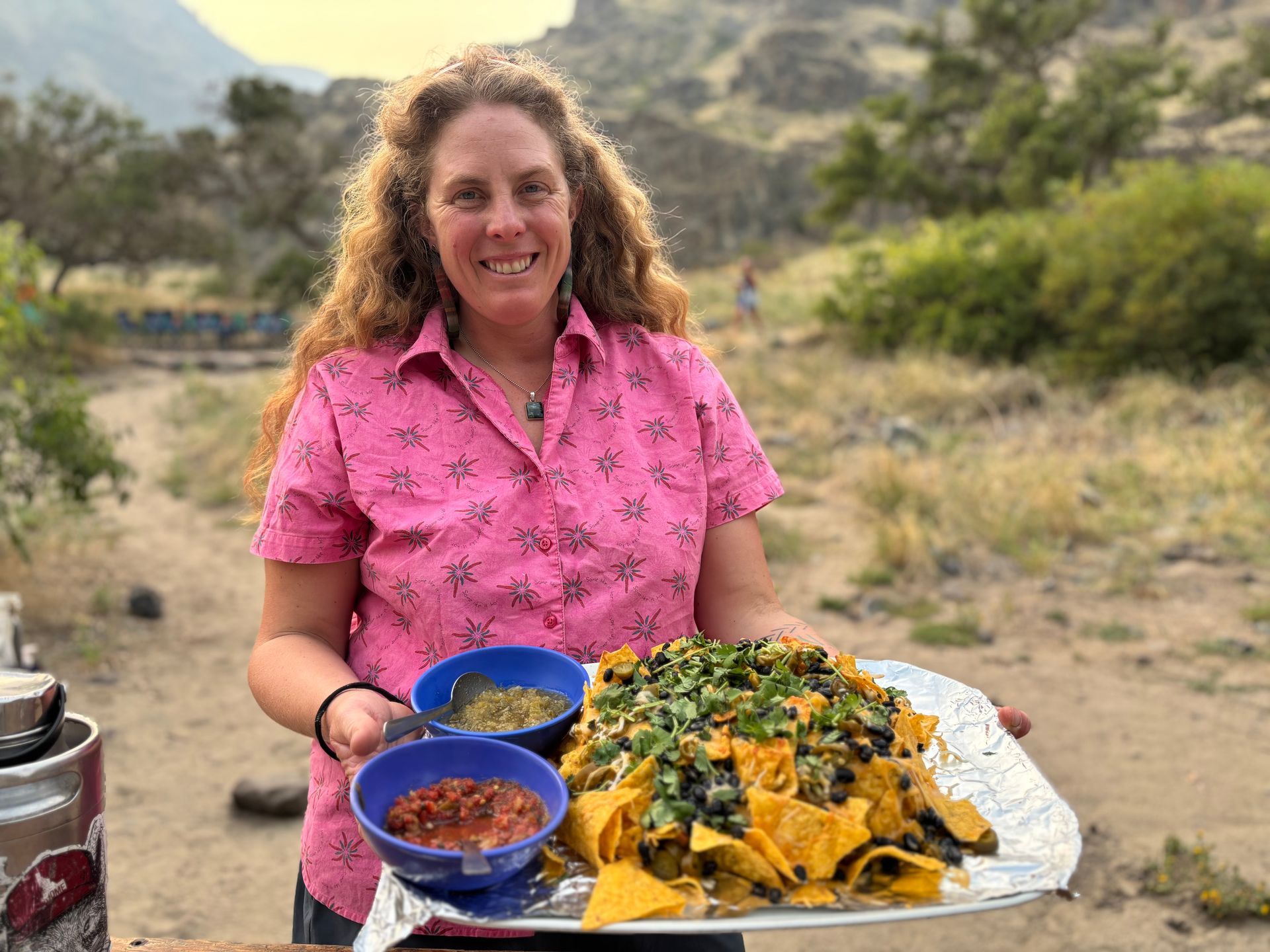 Woman holds tray of nachos with dips, smiling outdoors.