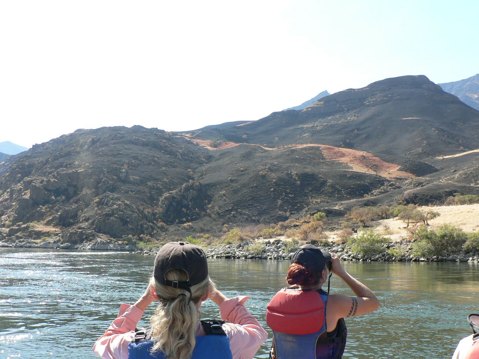 Two people in life jackets are looking out over a river with mountains in the background