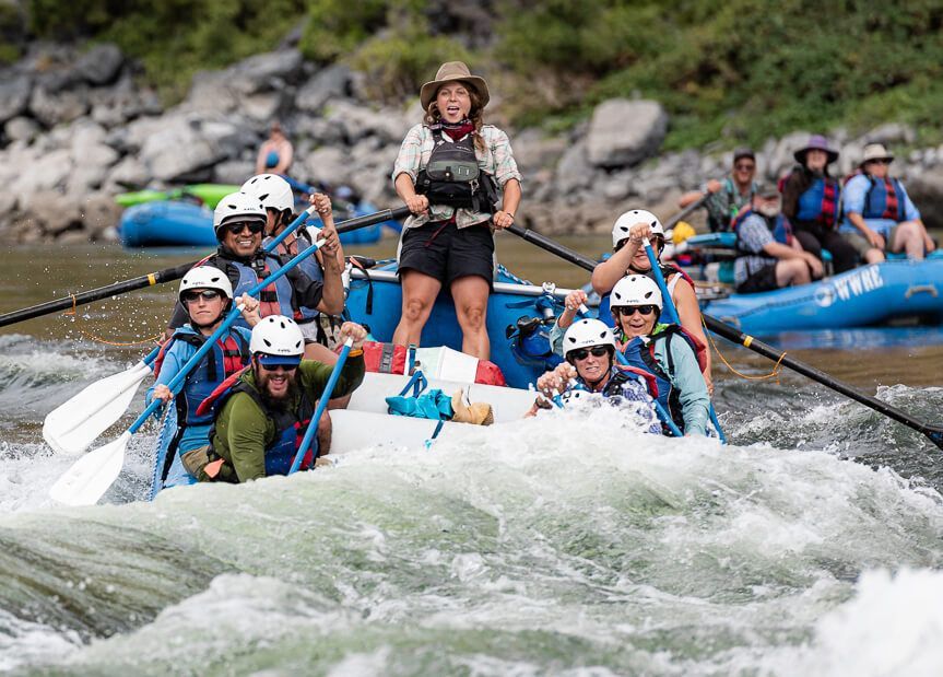 Rafting group on a whitewater river; people in helmets and life vests paddle, with one person standing.