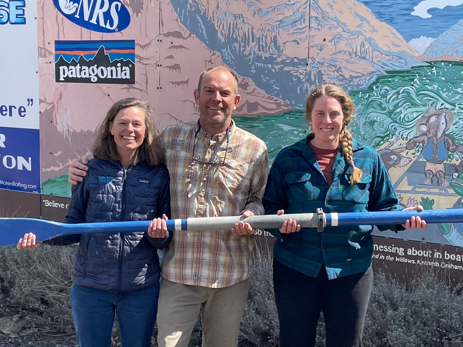 Three people hold a large blue oar in front of a mural. They stand outside on a sunny day.
