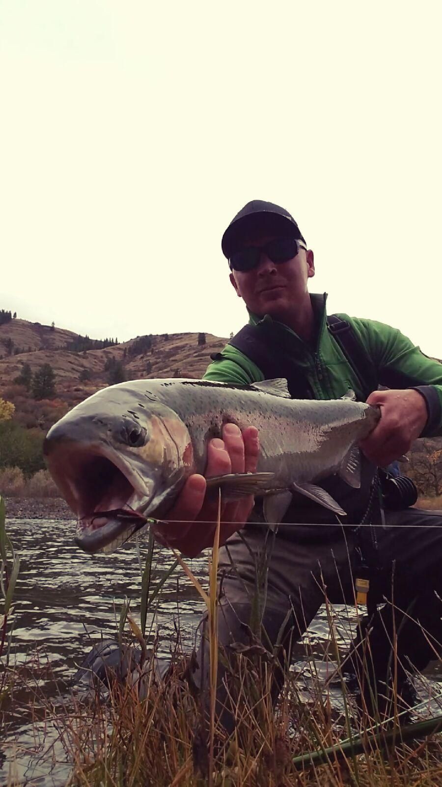Man holding large fish with open mouth near water and hills.