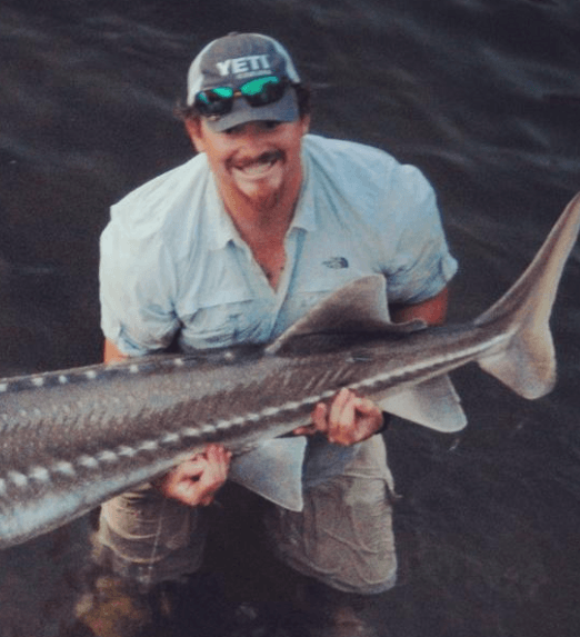 Man holding a large fish in water, smiling. He wears a Yeti hat, sunglasses, and light-colored clothing.