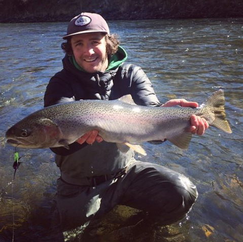 Man holding large rainbow trout in a river, smiling.