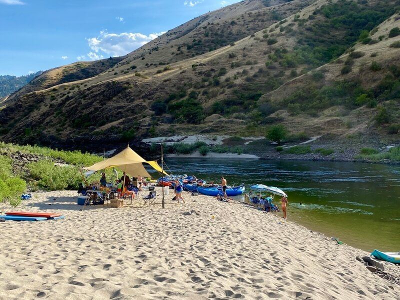 Group of People Are Sitting on A Sandy Beach Next to A River