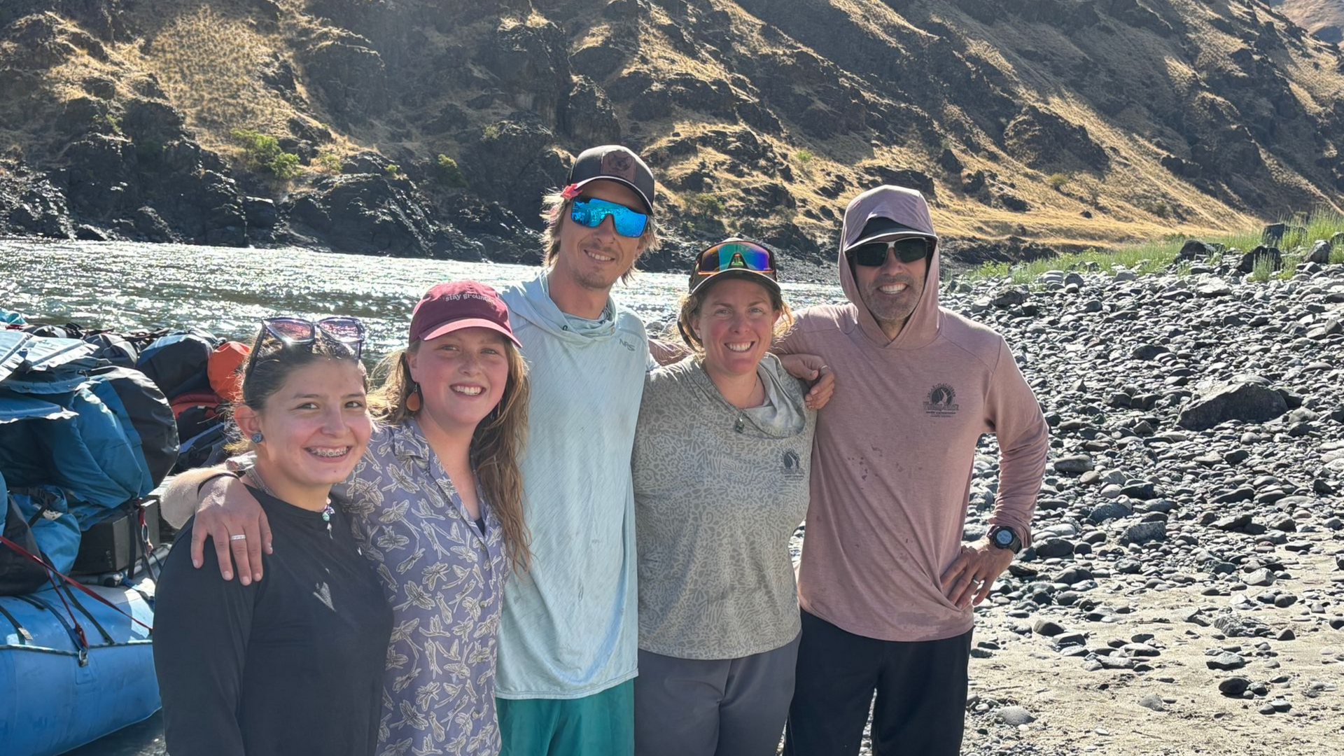Group of five people pose on a rocky shore next to a river. Mountains in the background.