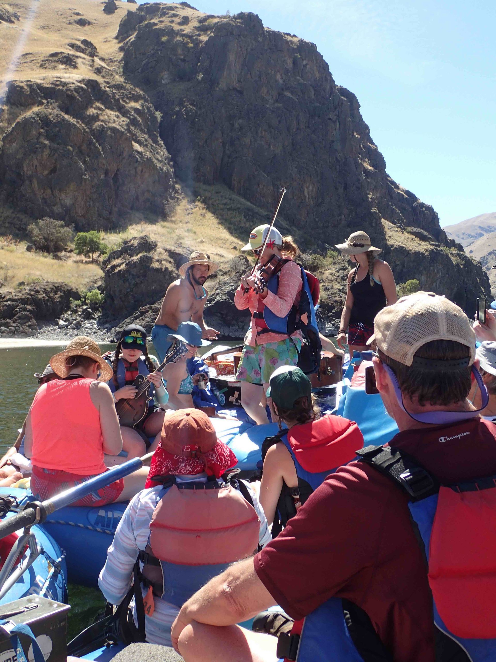 People on a raft in a river, rocky cliff in the background. Many wearing life vests, sunny day.
